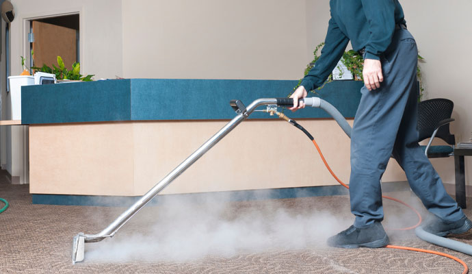 A professional cleaner uses a commercial steam cleaning wand to deep clean a carpet in an office lobby, with steam rising from the floor
