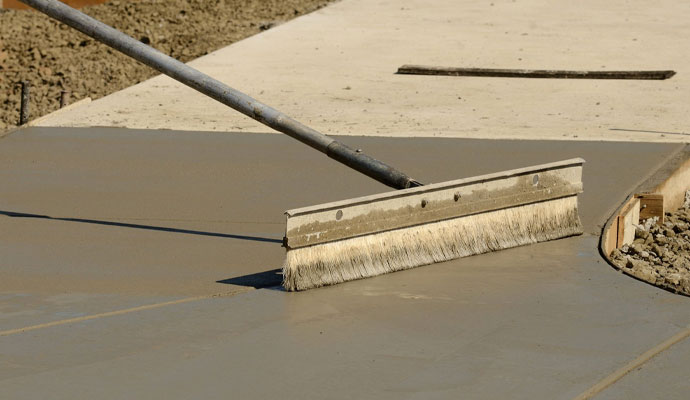 A close-up of a worker using a specialized broom to create a non-slip texture on a freshly poured concrete sidewalk or pathway