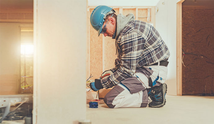 A professional electrician in a hard hat, safety glasses, and work clothes kneels on the floor, installing new electrical wiring in a house under construction