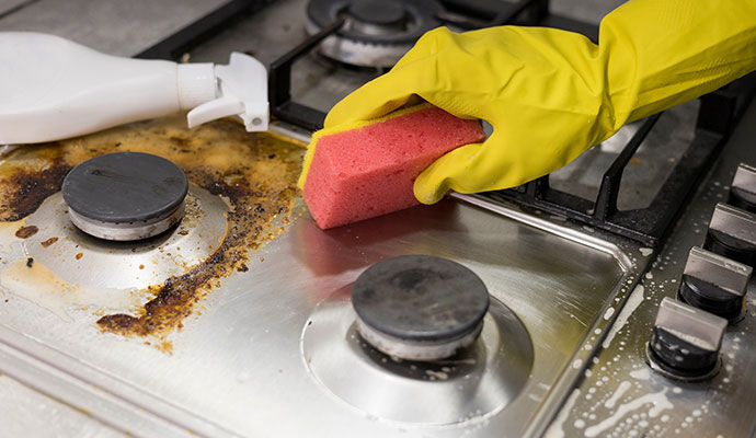 A person wearing a yellow glove uses a pink sponge and a cleaning solution to scrub a very greasy and dirty stainless steel gas stovetop