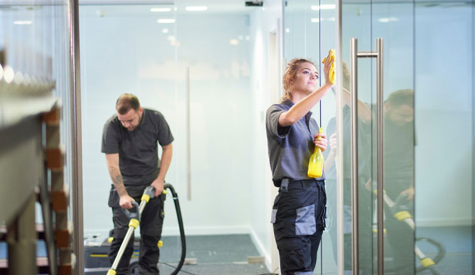 A man and a woman in work uniforms clean a modern office with large glass windows and carpeted floors