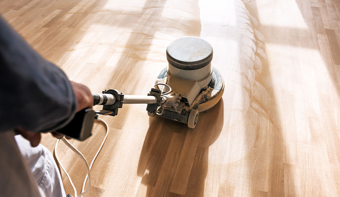 A professional uses a large sanding machine to refinish a wood floor, with saw marks and dust visible on the surface