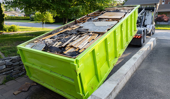 A large, bright green skip bin filled with junk and construction waste is placed on the curb of a suburban street