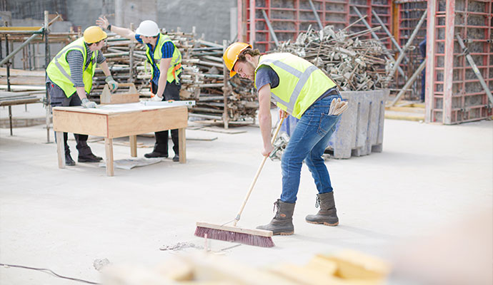 Construction workers on a job site, with one person sweeping the floor while two others discuss plans over a wooden table