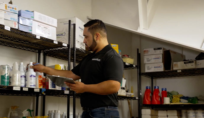 A REDLEE/SCS INC. employee in a black polo shirt holds a clipboard and reaches for a bottle on a shelf while taking inventory in a stockroom filled with cleaning supplies