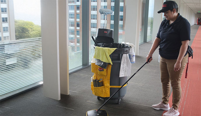 A professional cleaner in a baseball cap and uniform uses a mop in an office hallway next to a large, fully-stocked cleaning cart