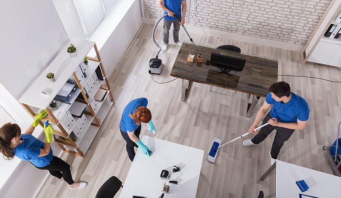 Group of janitorial staff members cleaning an office, one vacuuming, one mopping and another dusting a shelf