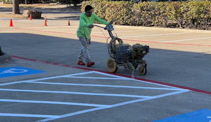 A person in a green shirt operates a line striping machine to paint white lines on a commercial parking lot, with a blue handicap symbol visible nearby