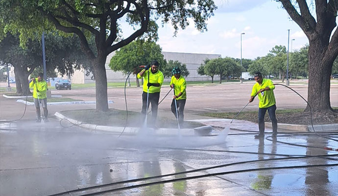 A team of four workers in bright yellow safety vests uses high-pressure sprayers to clean a parking lot, with water splashing on the pavement