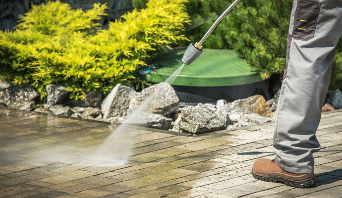 A person in work boots and pants uses a pressure washer to clean a tiled outdoor walkway or patio next to a rock garden and bushes