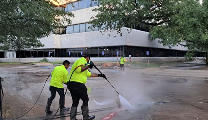 A team of professional cleaners in bright yellow safety vests and rubber boots uses high-pressure water sprayers to clean a dirty commercial parking lot