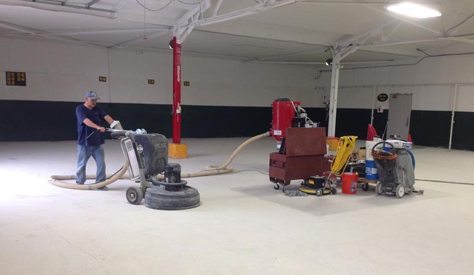 A person operates a large floor grinder with a connected vacuum hose in an empty warehouse or commercial space